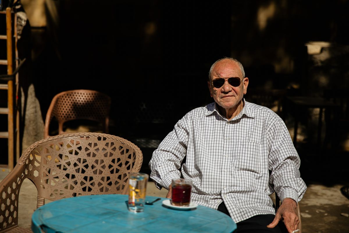An elderly man sitting at an outdoor café table in the morning sun.