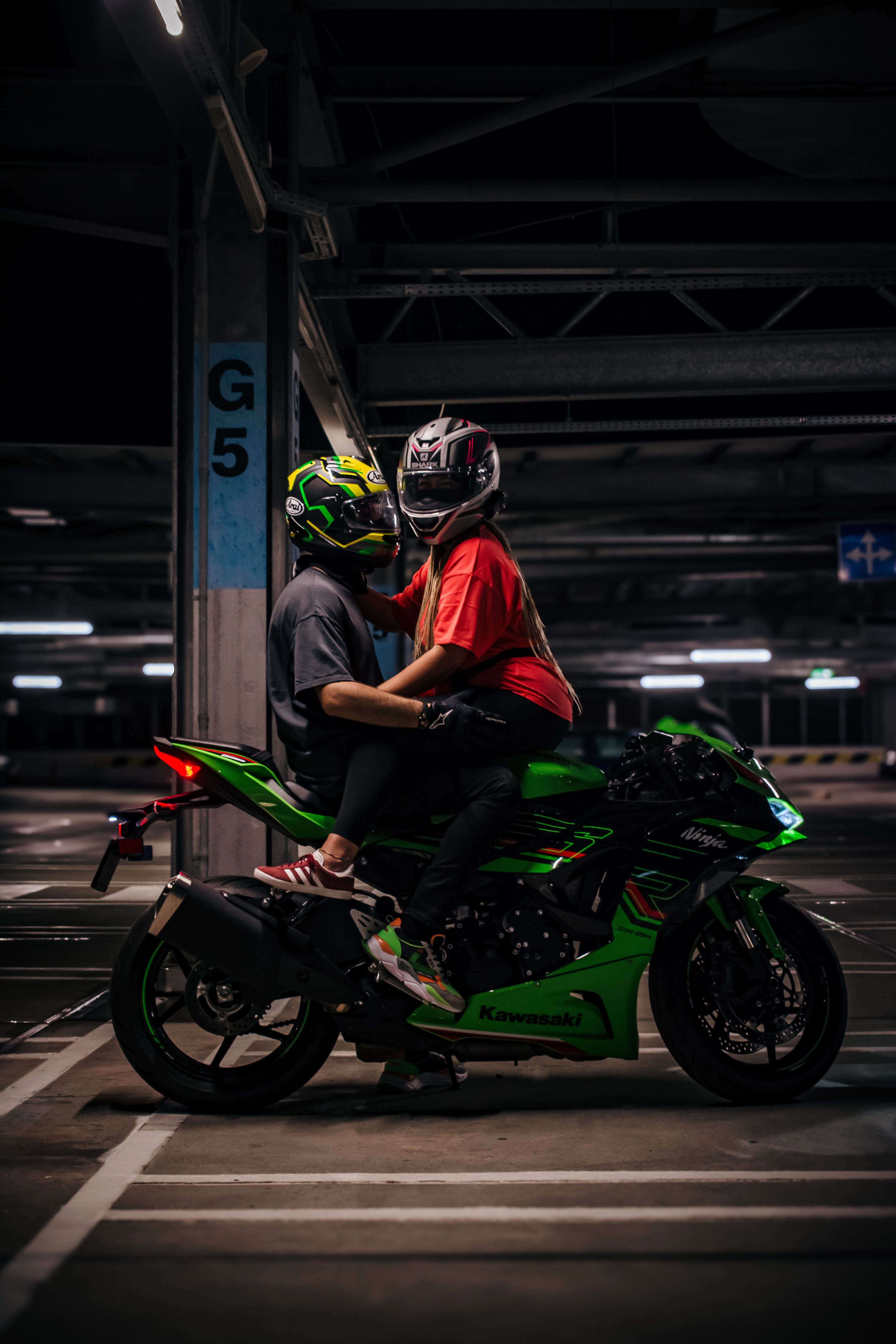 A couple in helmets on a bright green motorcycle in a dimly lit parking garage.