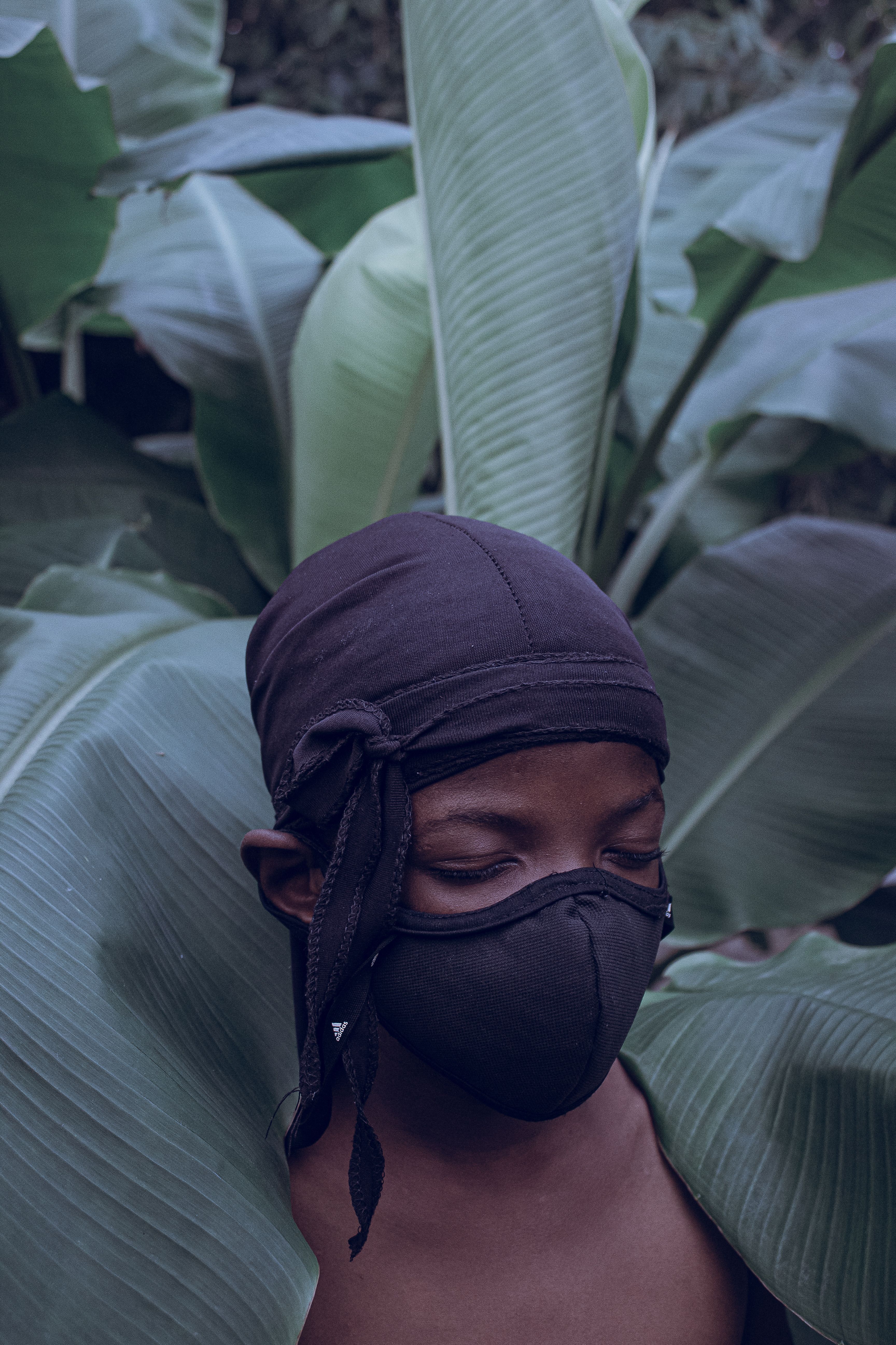 Person wearing a mask amidst lush green leaves, evoking nature and mystery.