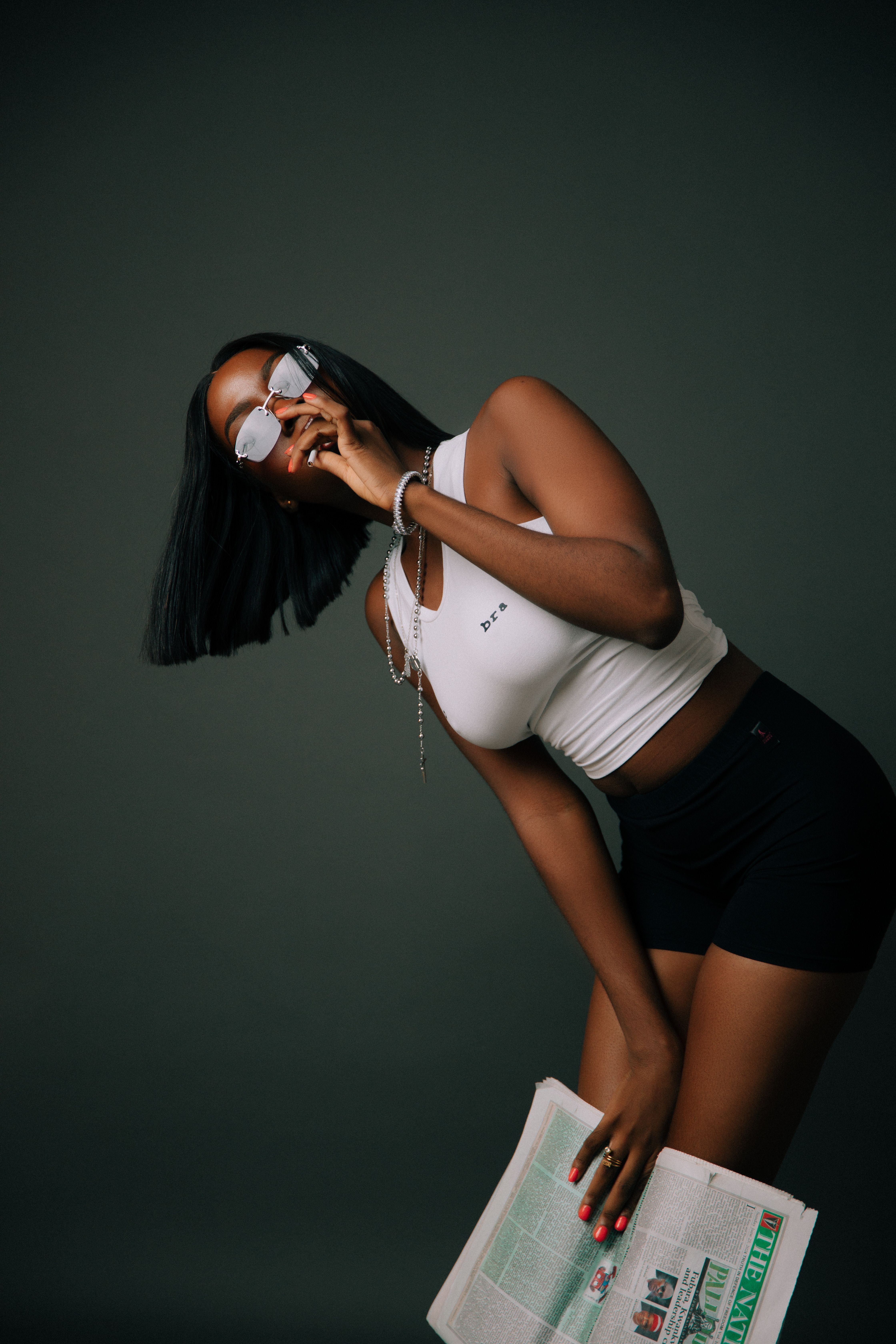 Fashionable young woman in sunglasses poses with a newspaper indoors.