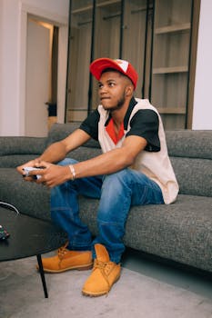A young man intensely playing video games on a sofa, wearing casual attire and a red cap.