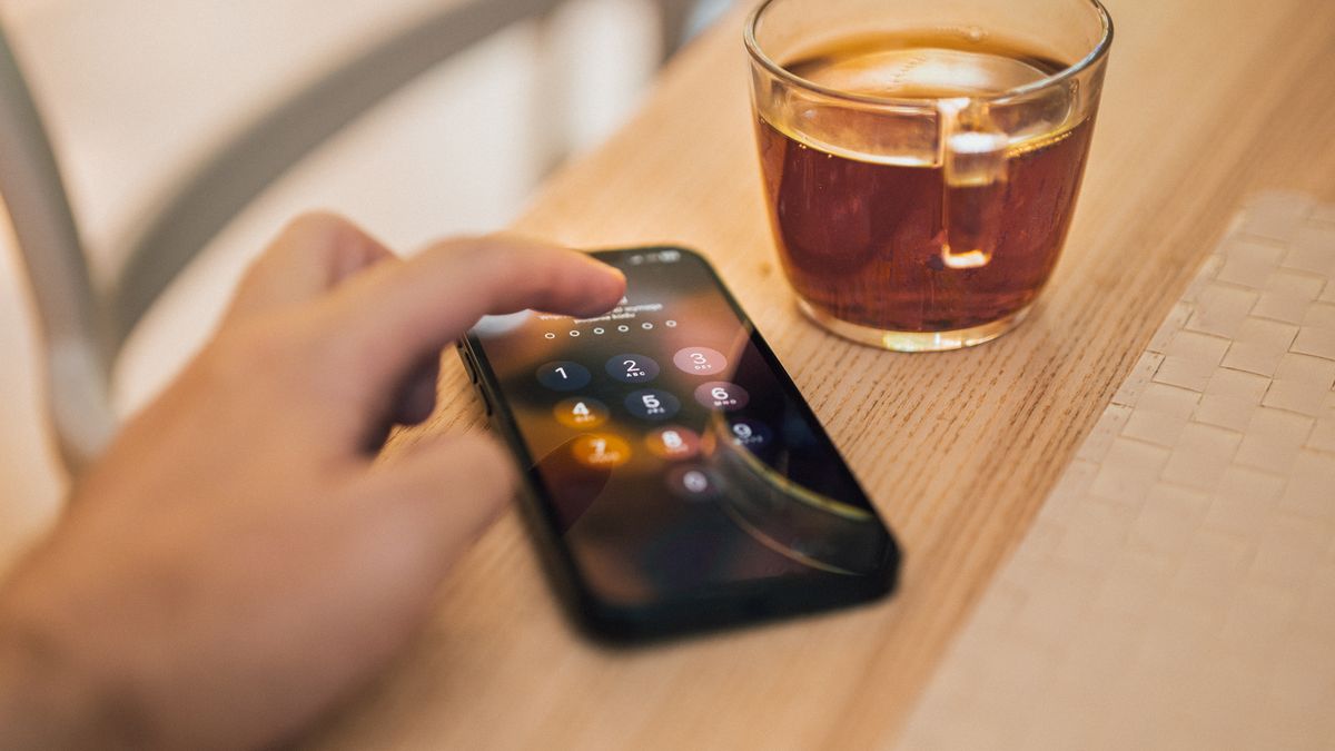 Mão desbloqueando a tela de um smartphone sobre uma mesa de madeira, ao lado de uma xícara