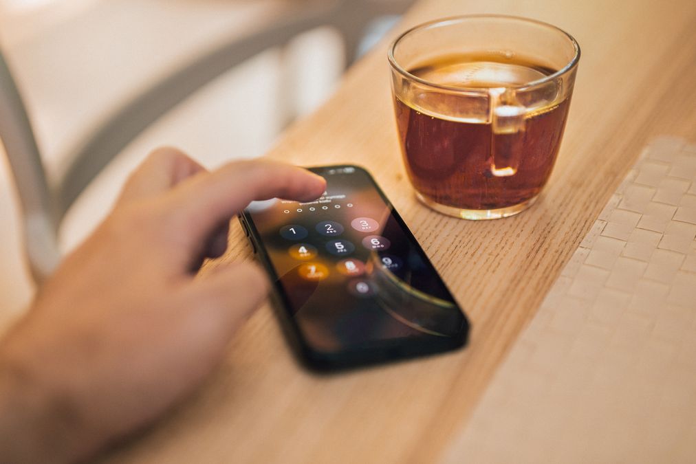 Mão desbloqueando a tela de um smartphone sobre uma mesa de madeira, ao lado de uma xícara