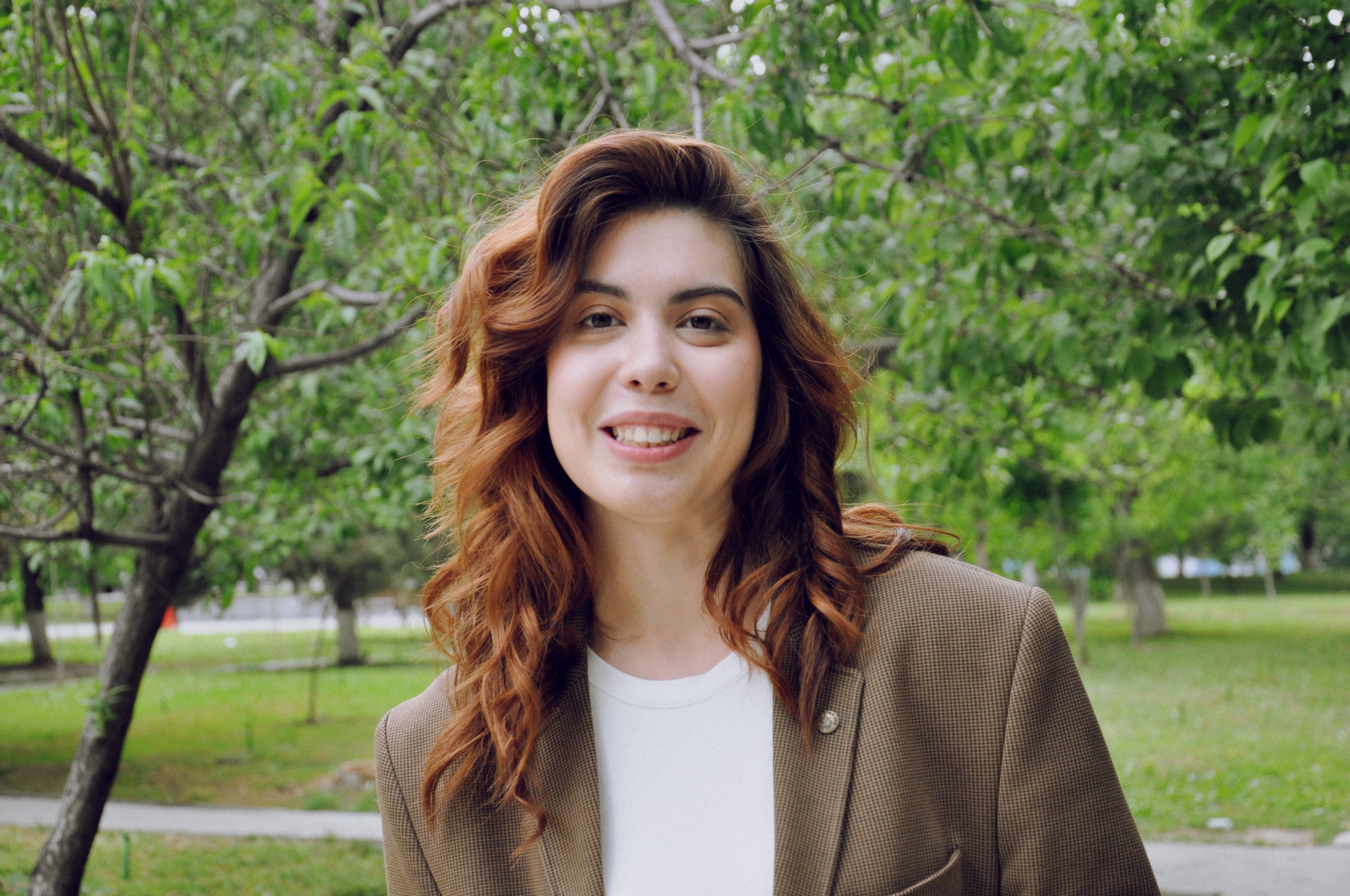 A woman with curly brown hair smiles outdoors in a lush, green park.