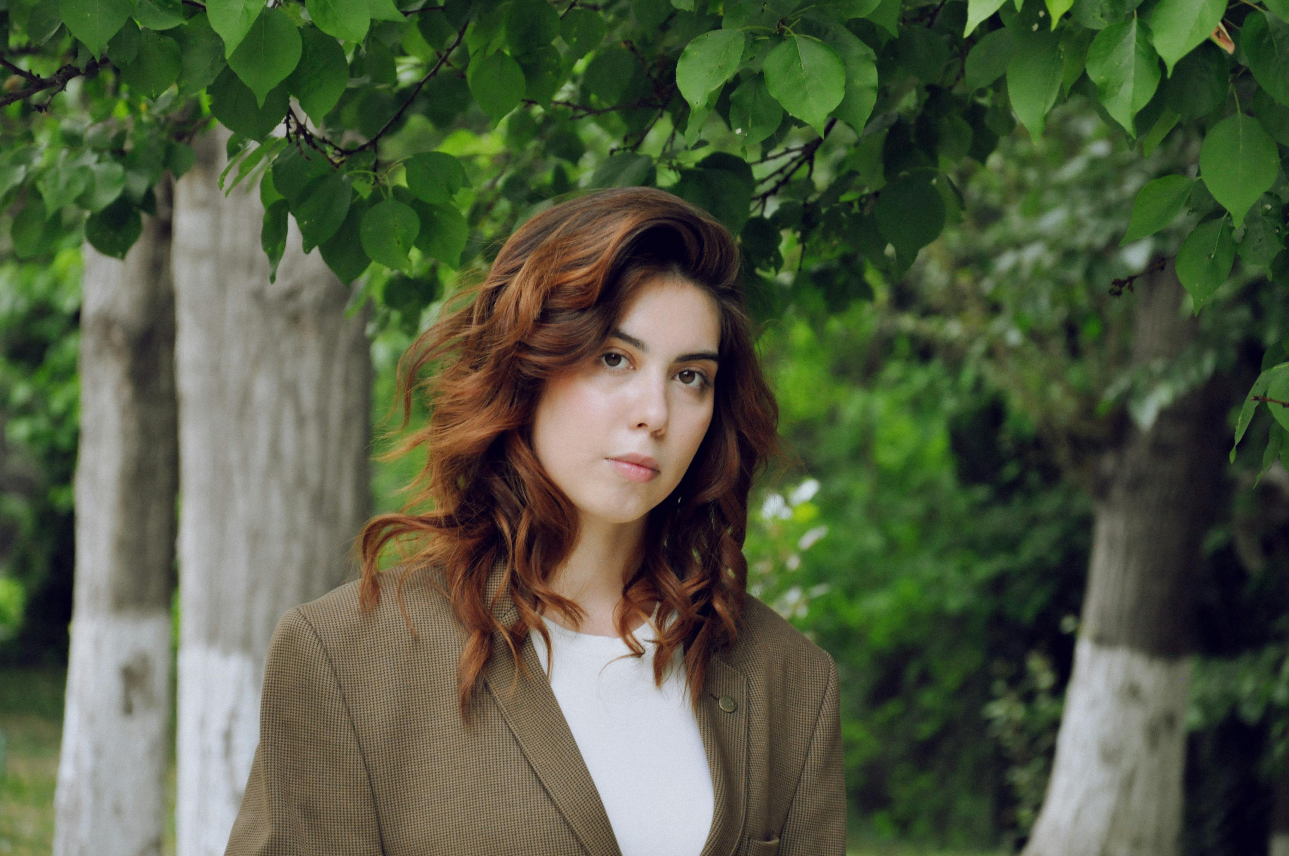Free A woman with wavy hair stands in a lush green park, wearing a brown blazer. Stock Photo
