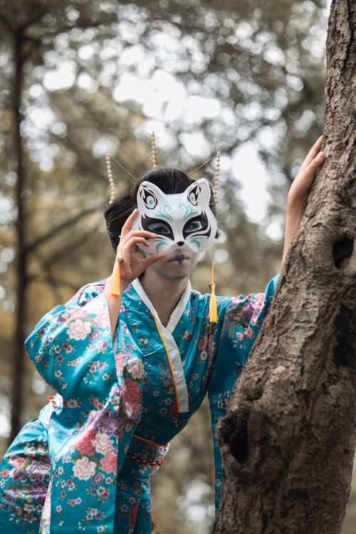 Free Woman in a floral kimono wearing a kitsune mask posing by a tree in a park. Stock Photo
