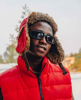 Portrait of a stylish man wearing sunglasses, a red vest, and a fur-lined hat outdoors.