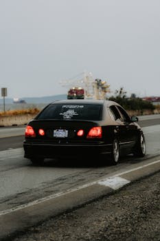 Black car speeding on a coastal highway with a hazy industrial backdrop, captured in motion.