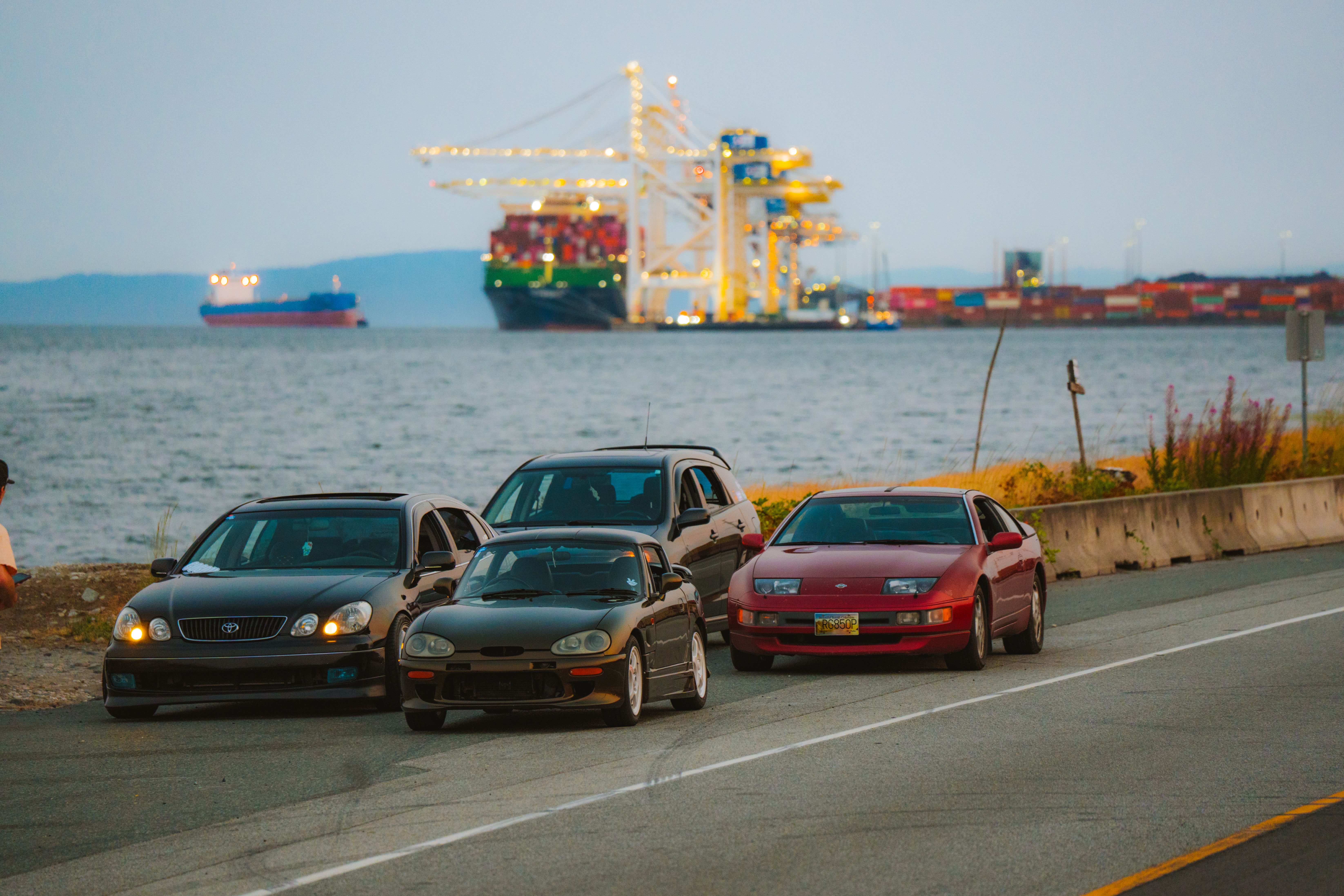Three parked cars by the seashore with cargo ships and cranes at a harbor nearby, during the day.