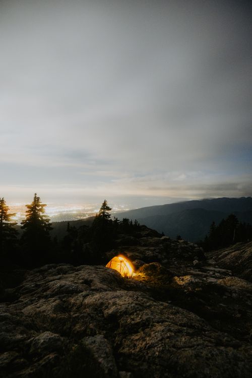Free A peaceful camping scene at night with a glowing tent amidst a mountainous landscape under a cloudy sky. Stock Photo