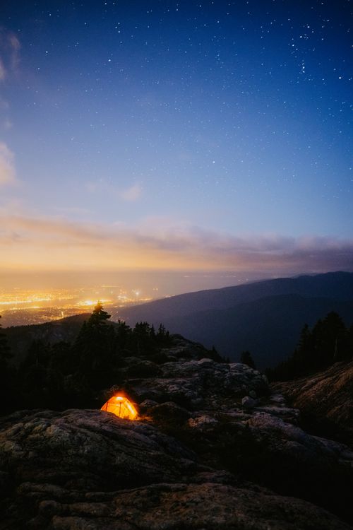 Free A mountain camping scene with a glowing tent under a starlit sky overlooking a cityscape. Stock Photo