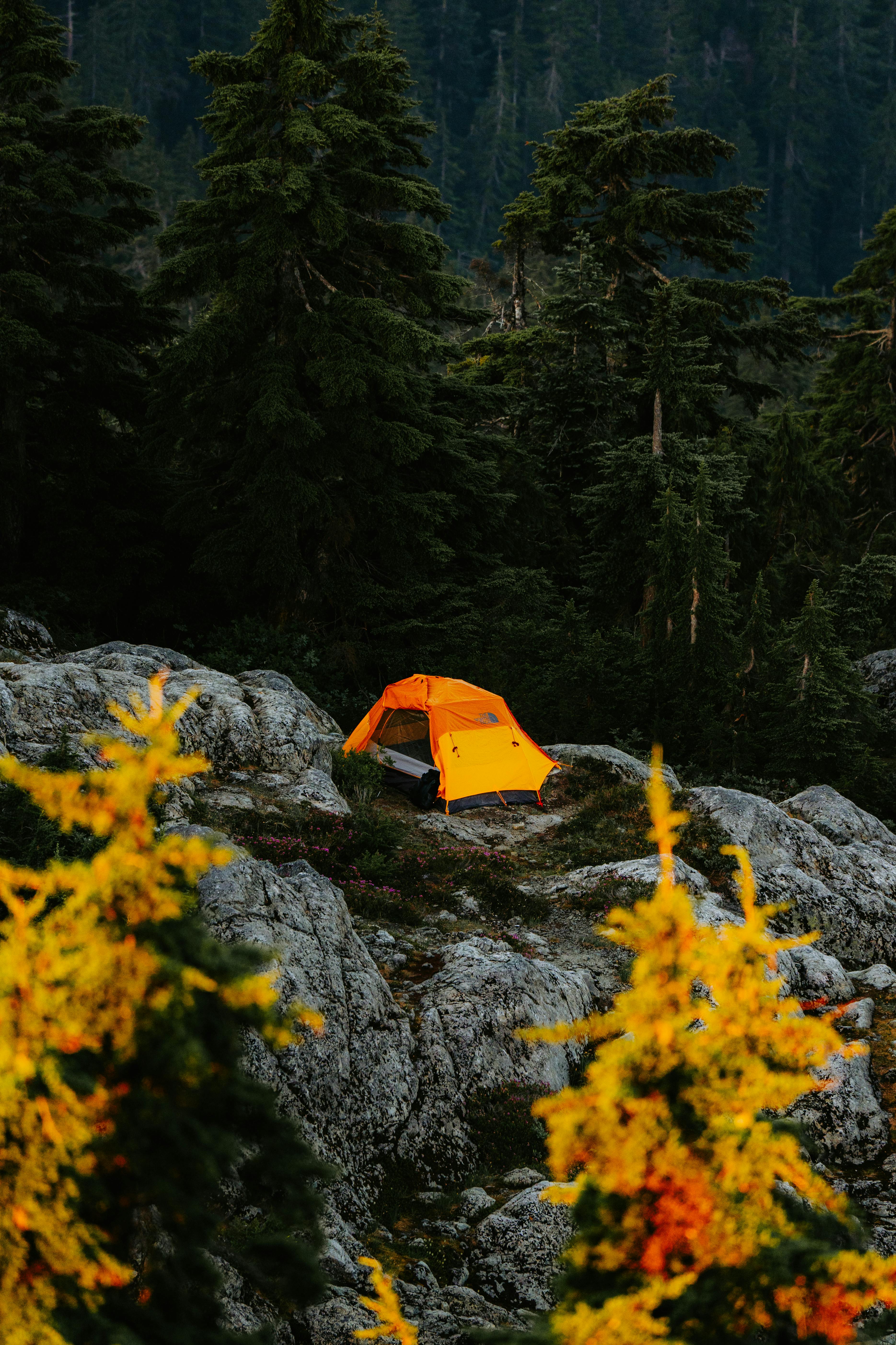 A bright orange tent set amidst a rocky mountain terrain with lush green trees, capturing autumn's beauty.