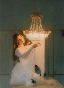 A serene woman in a white gown delicately adjusts a chandelier in a vintage room.