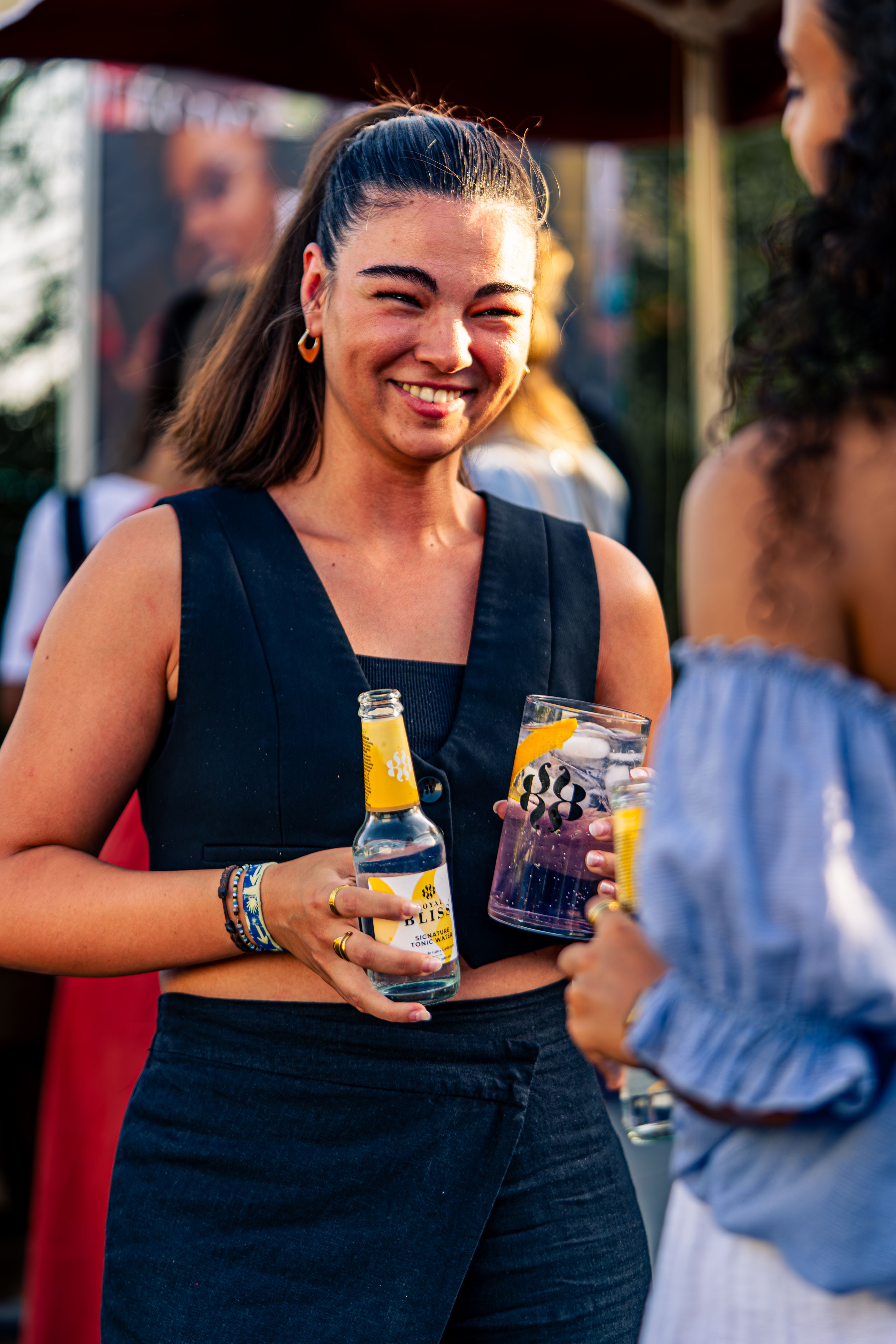 Smiling woman holding drinks at an outdoor summer festival, enjoying time with friends.