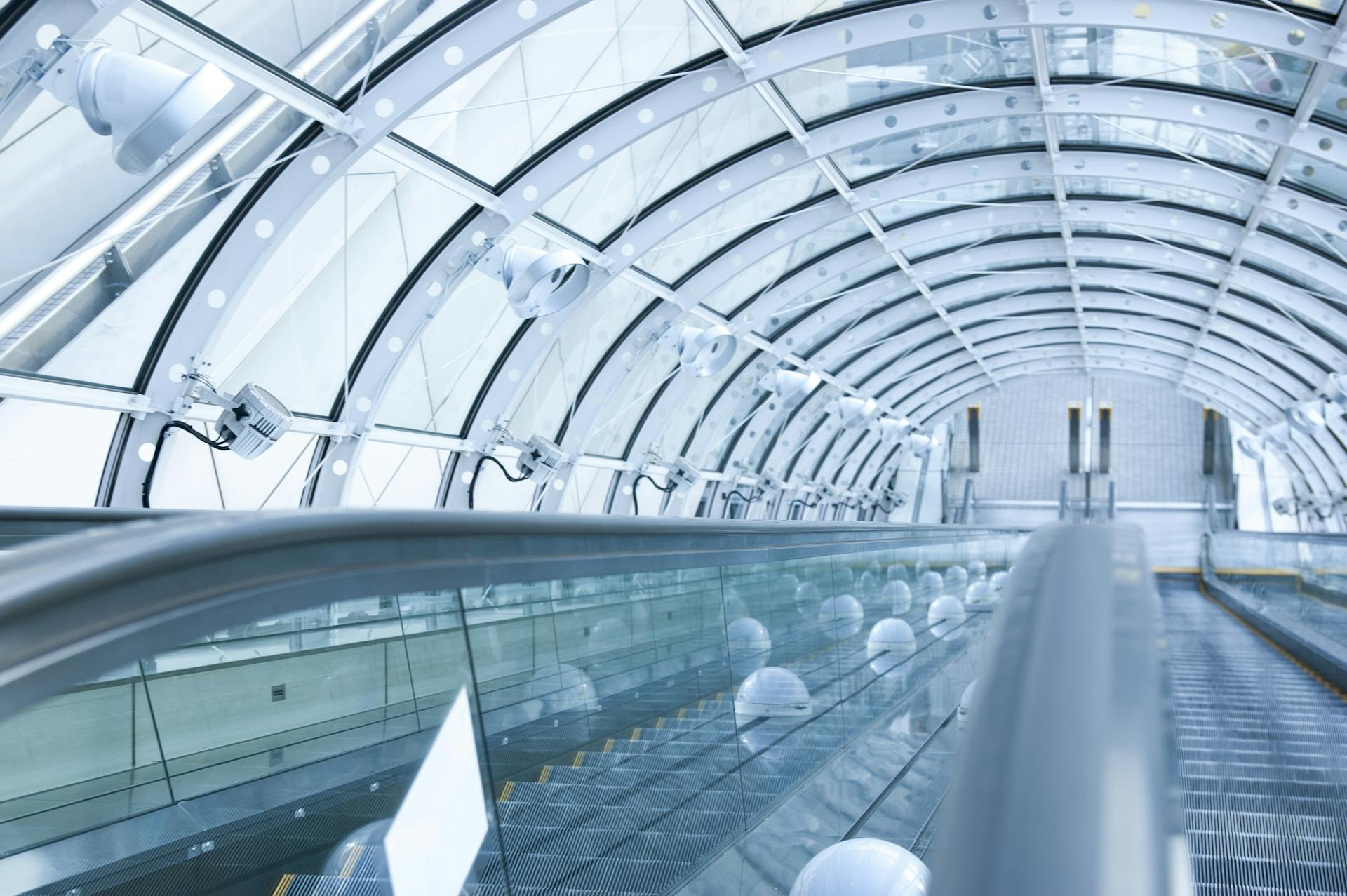 Futuristic glass tunnel interior featuring an escalator with steel and glass architecture.