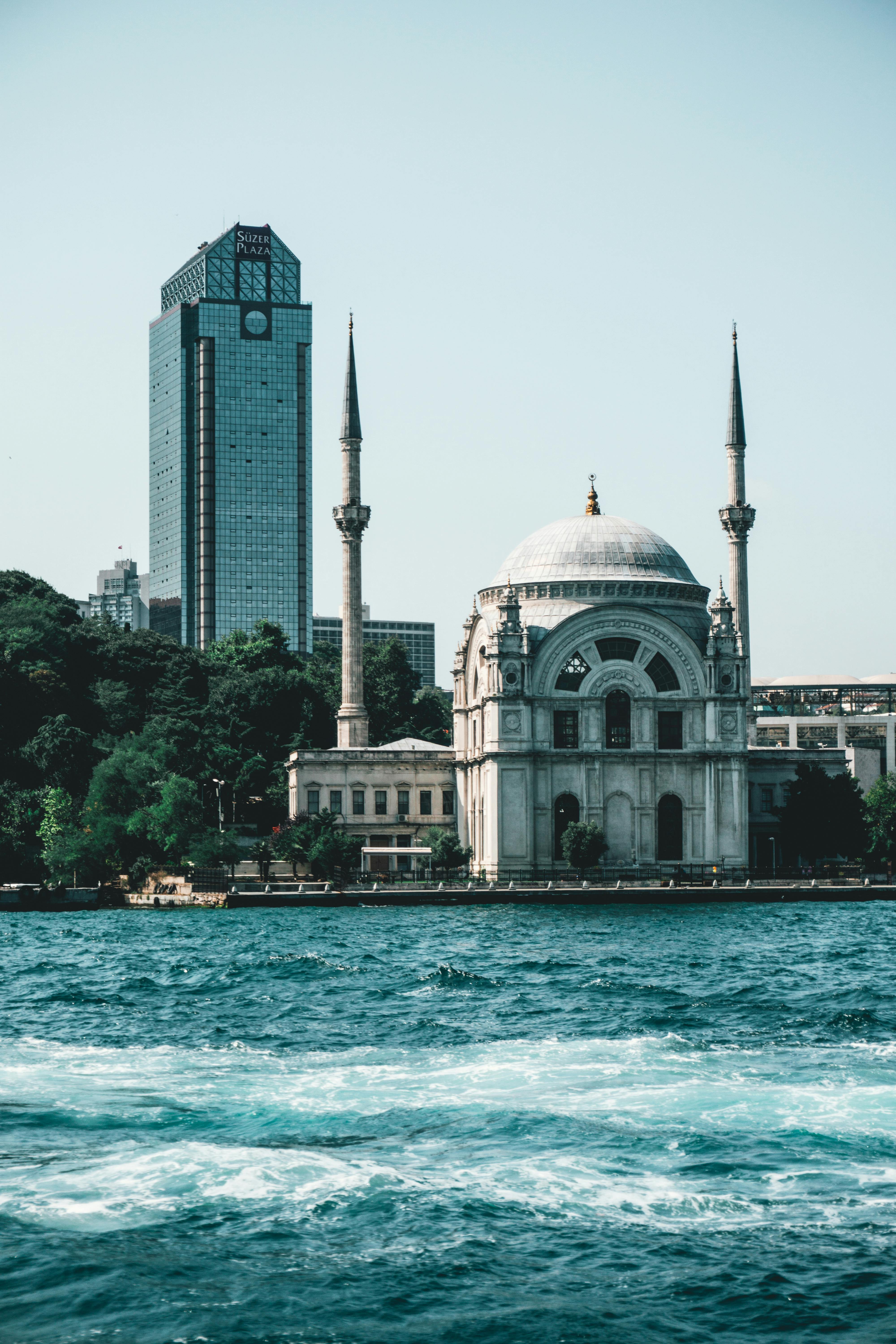 在晴朗的天空下，历史悠久的清真寺与现代城市的天际线在水边构成了一幅风景