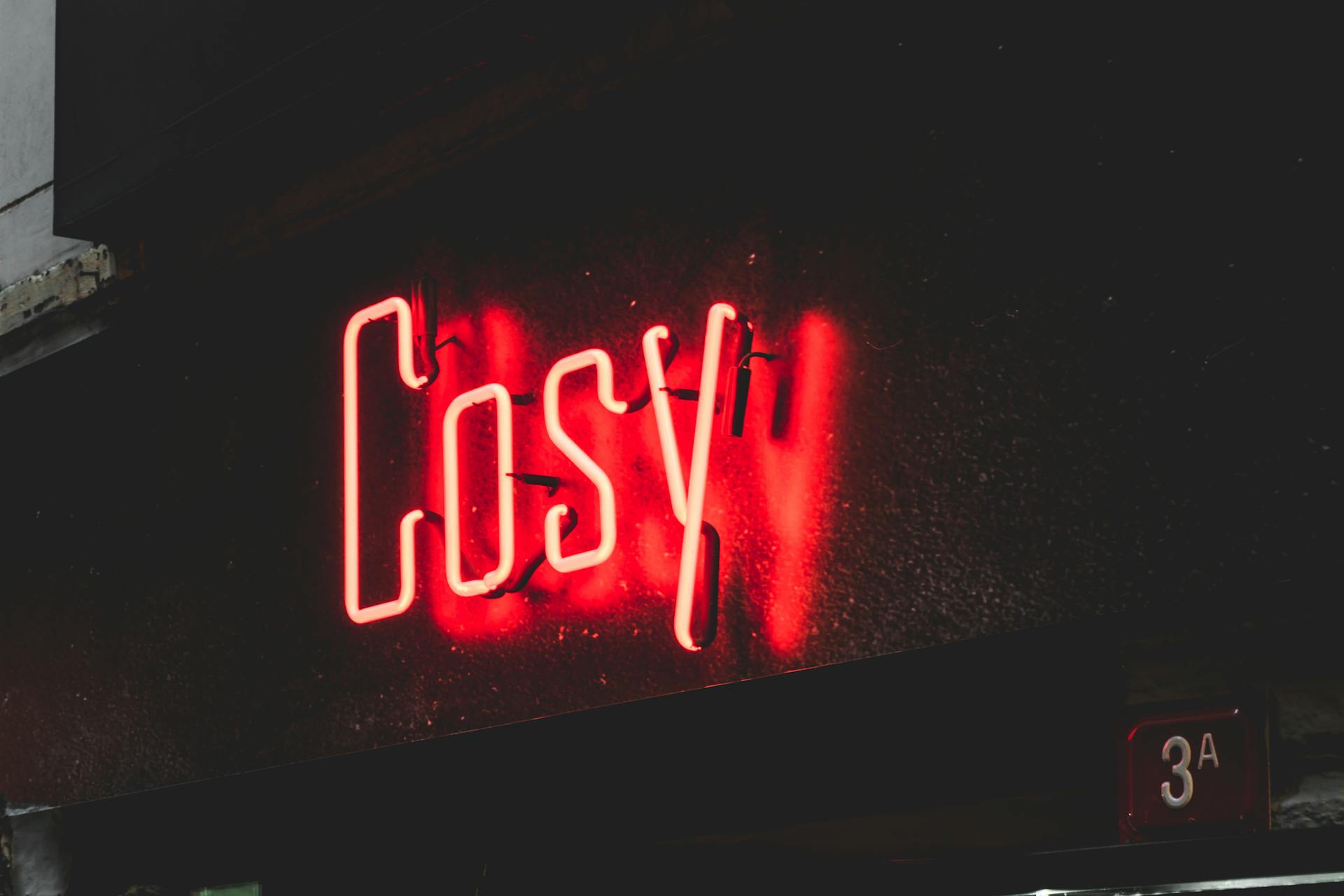 Glowing red neon 'Cosy' sign illuminating a dark urban street corner at night.