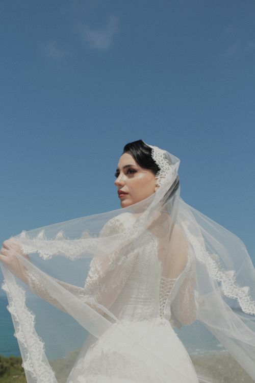 Free An elegant bride stands outdoors holding her veil against a clear blue sky, capturing wedding beauty. Stock Photo