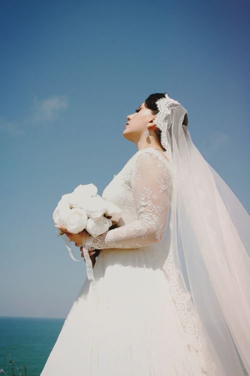 Free A bride in a lace dress and veil holds a bouquet by the ocean under a clear blue sky. Stock Photo