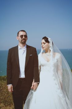 Bride and groom holding hands outdoors with ocean view, wearing elegant attire.