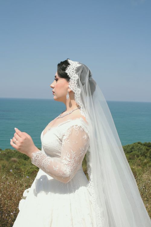 Free Elegant bride in lace dress and veil posing by the ocean under a clear blue sky. Stock Photo