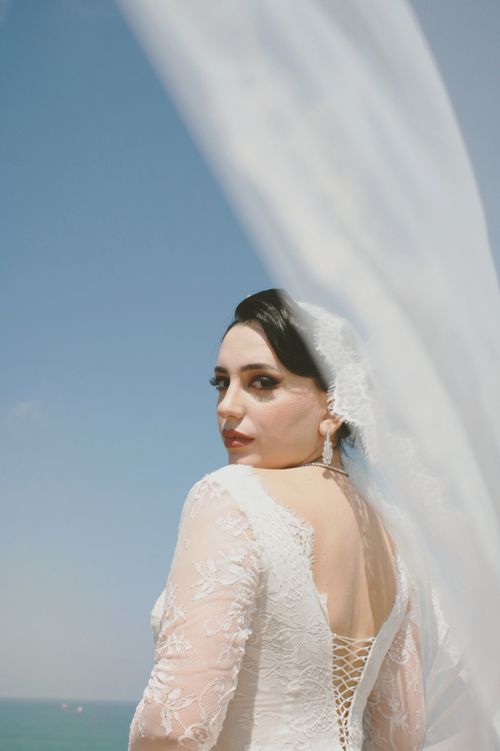 Free A bride in a lace dress with a flowing veil looking over her shoulder at the beach. Stock Photo
