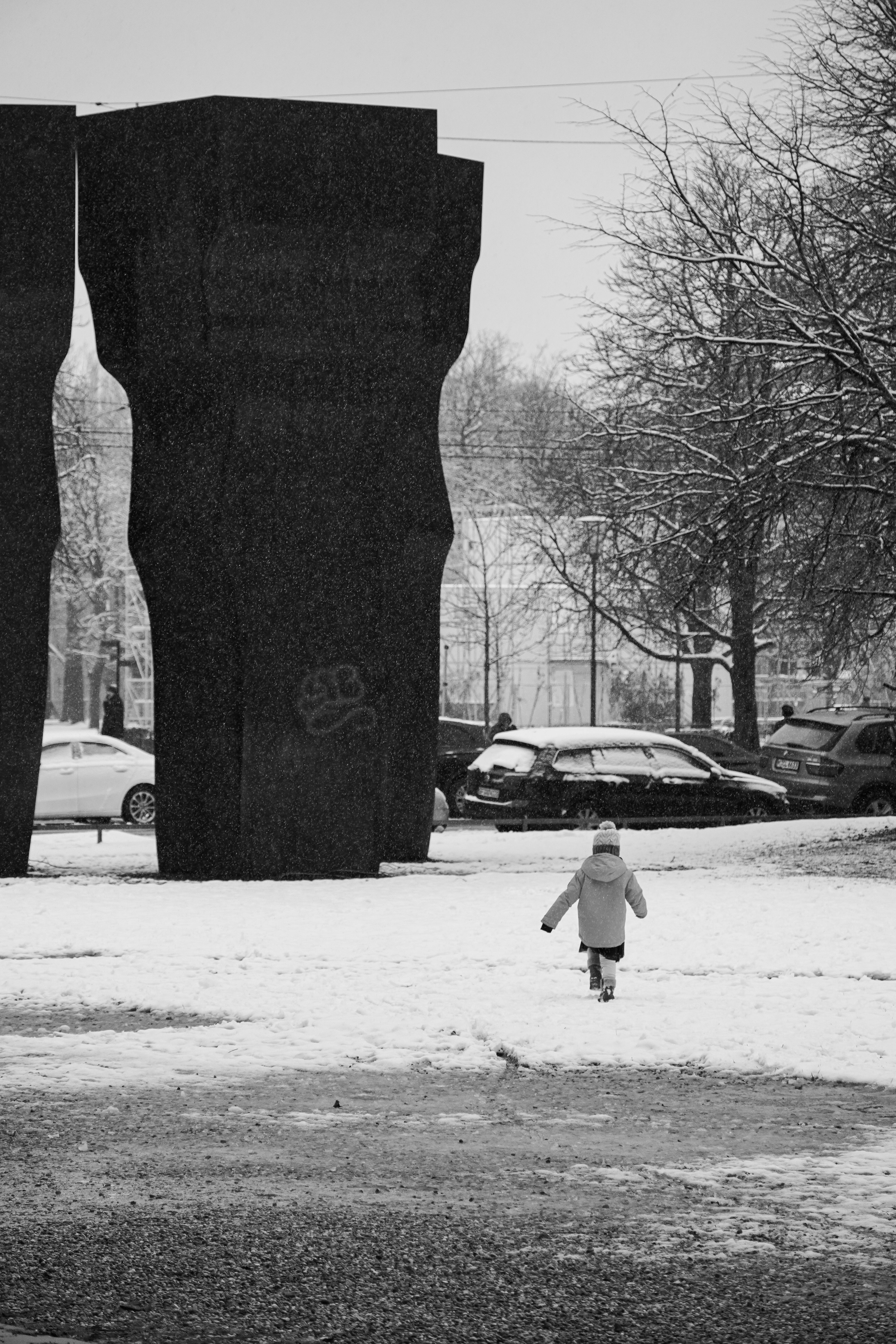A child in winter attire walks towards Eduardo Chillida sculptures in snowy Munich park.