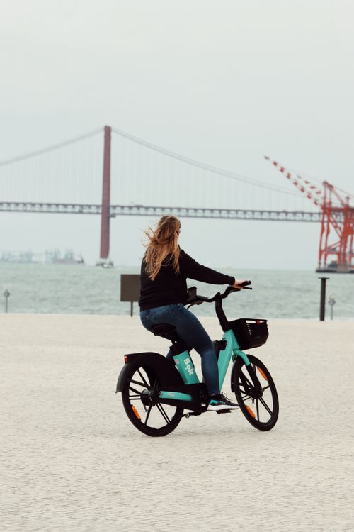 Free A woman rides a bike by the 25 de Abril Bridge in Lisbon, showing leisure travel. Stock Photo