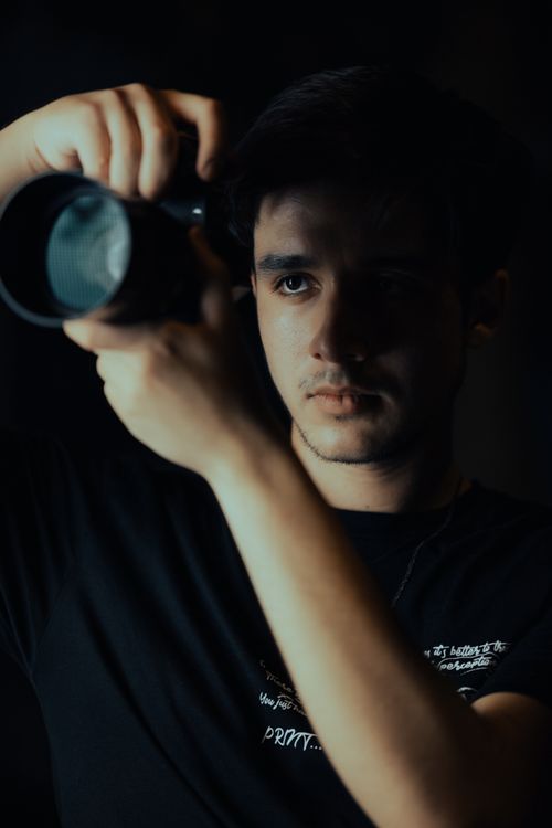 Free A portrait of a man holding a camera lens in a dimly lit studio, capturing focus and intensity. Stock Photo