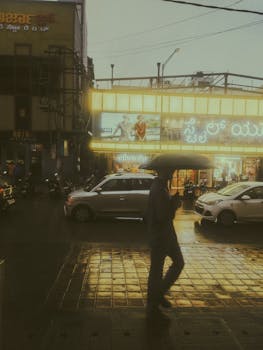 A person with an umbrella crossing a busy street in rainy urban India, with illuminated buildings and vehicles in the background.
