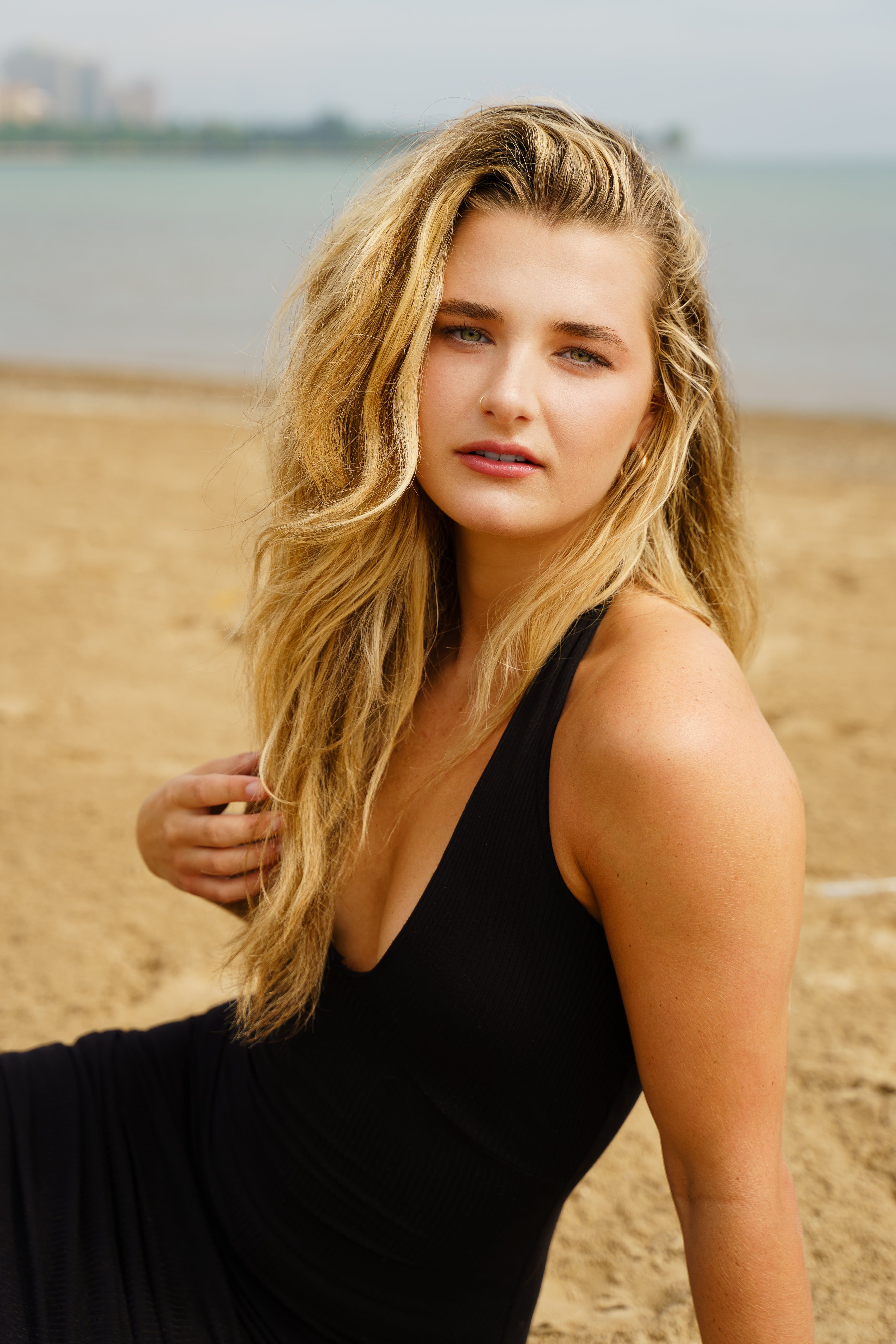 Elegant woman in a black dress sits on a Chicago beach, exuding sophistication and beauty.