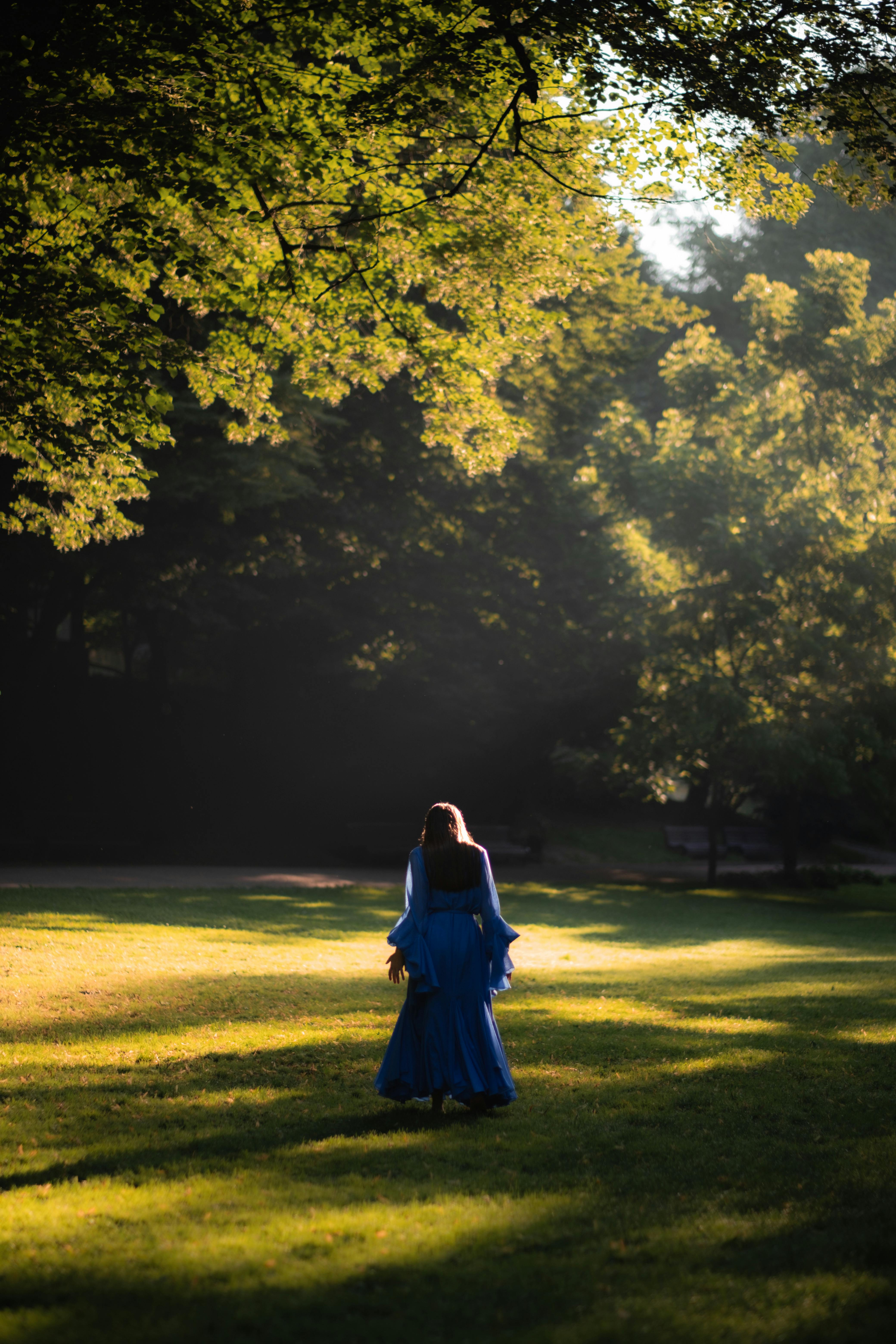 A woman in a flowing blue dress walks through a sunlit park surrounded by vibrant trees.