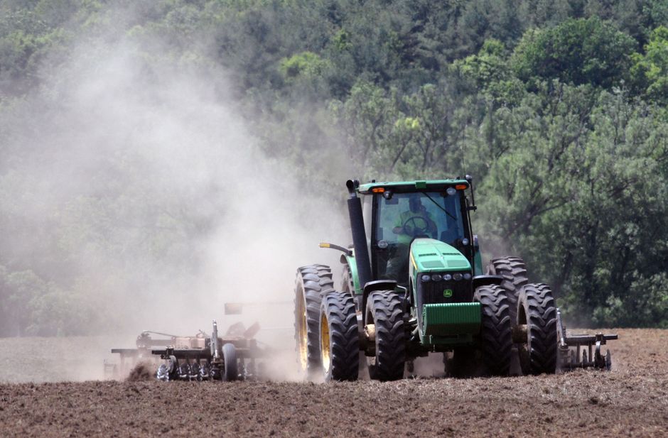 A green tractor plowing a field, creating clouds of dust in an agricultural setting.