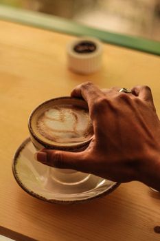 Close-up of a hand holding a latte with foam art on a saucer at a cozy cafe table.
