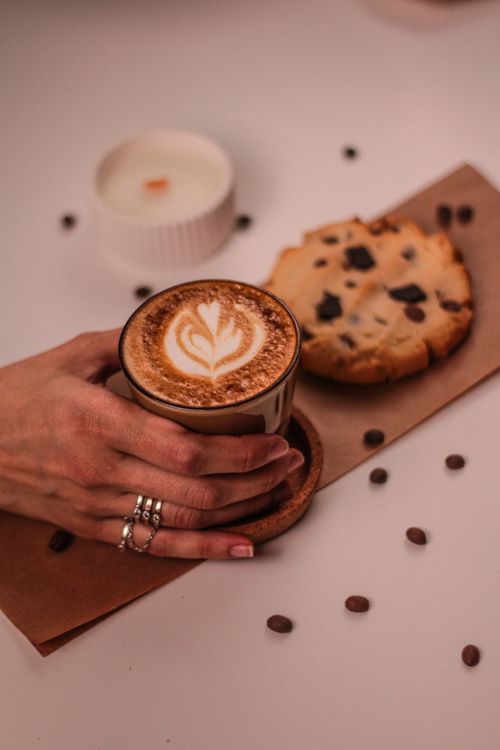 Free Aesthetic flat lay of cappuccino with latte art, chocolate chip cookie, and a candle on a wooden table. Stock Photo