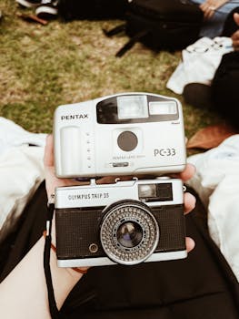 Two vintage analog cameras, Pentax and Olympus, held by a person outdoors on a grassy area.