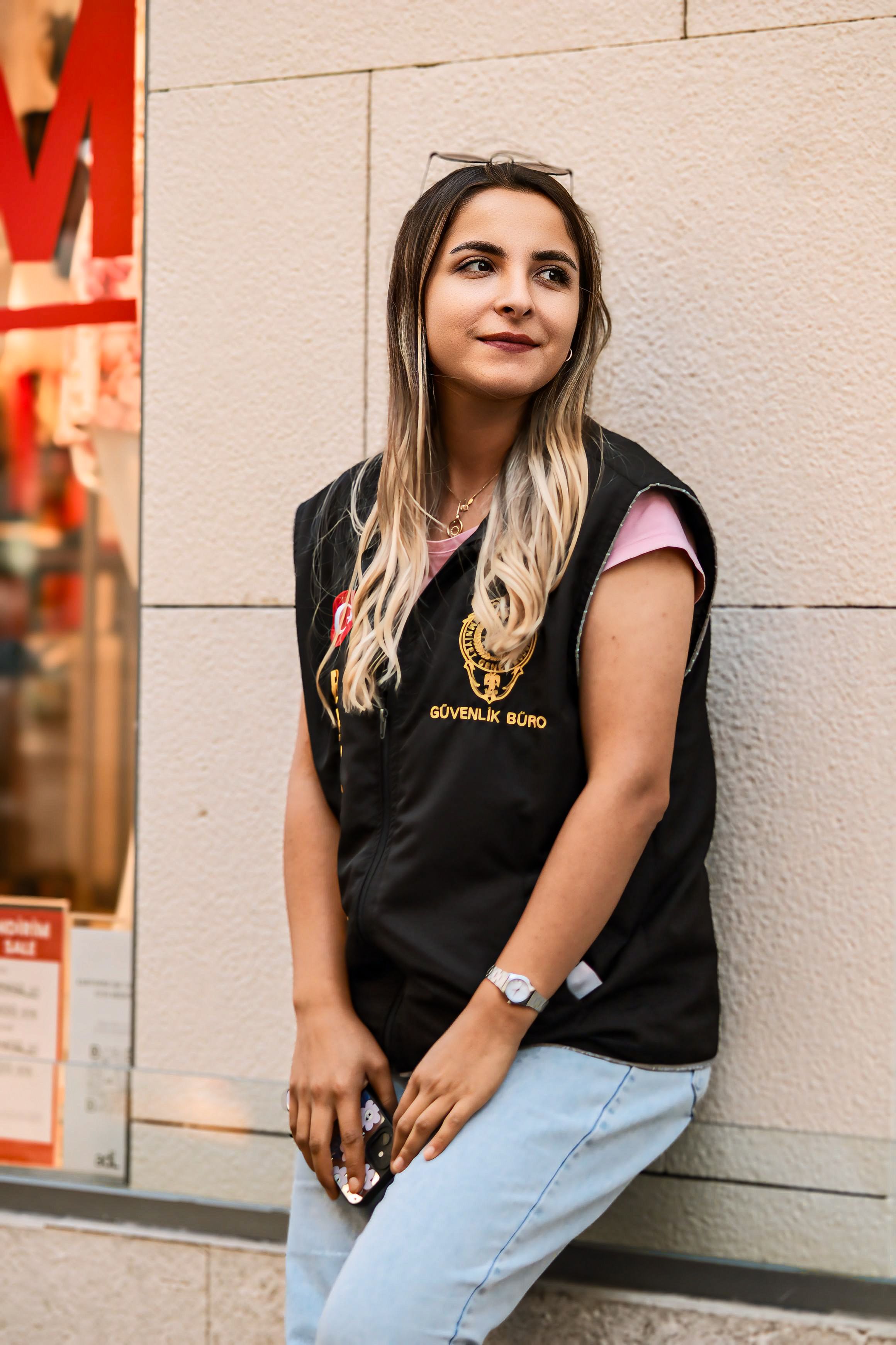 Stylish young woman with long hair leaning against a city wall, radiating confidence in urban fashion.