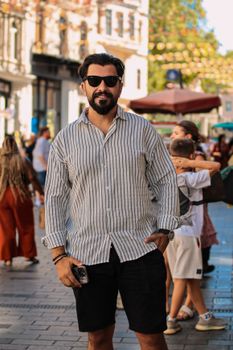 Fashionable man in sunglasses poses confidently on a lively city street during the day.