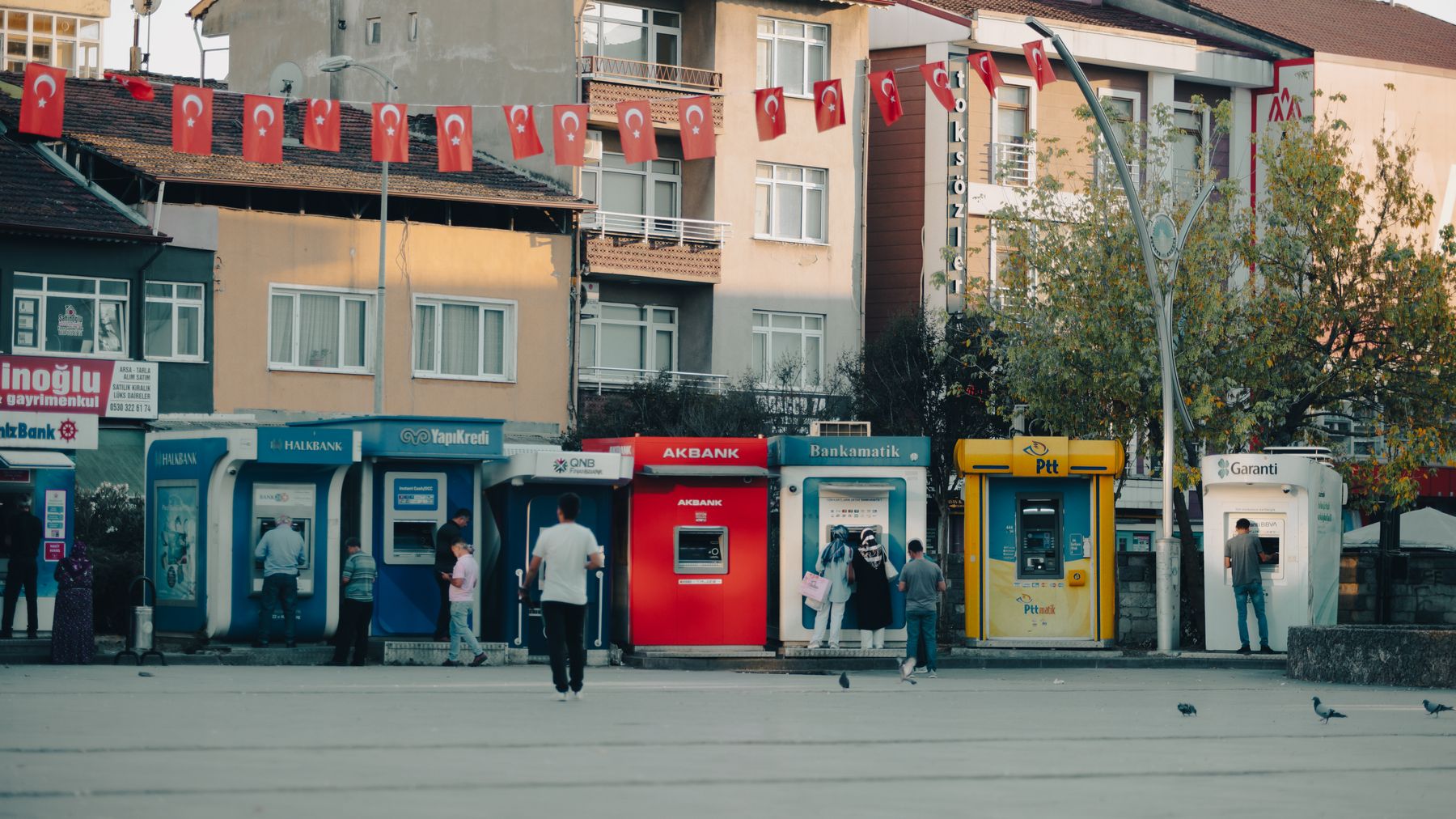 Street view in Bartin, Turkiye with ATMs, people, and waving Turkish flags.