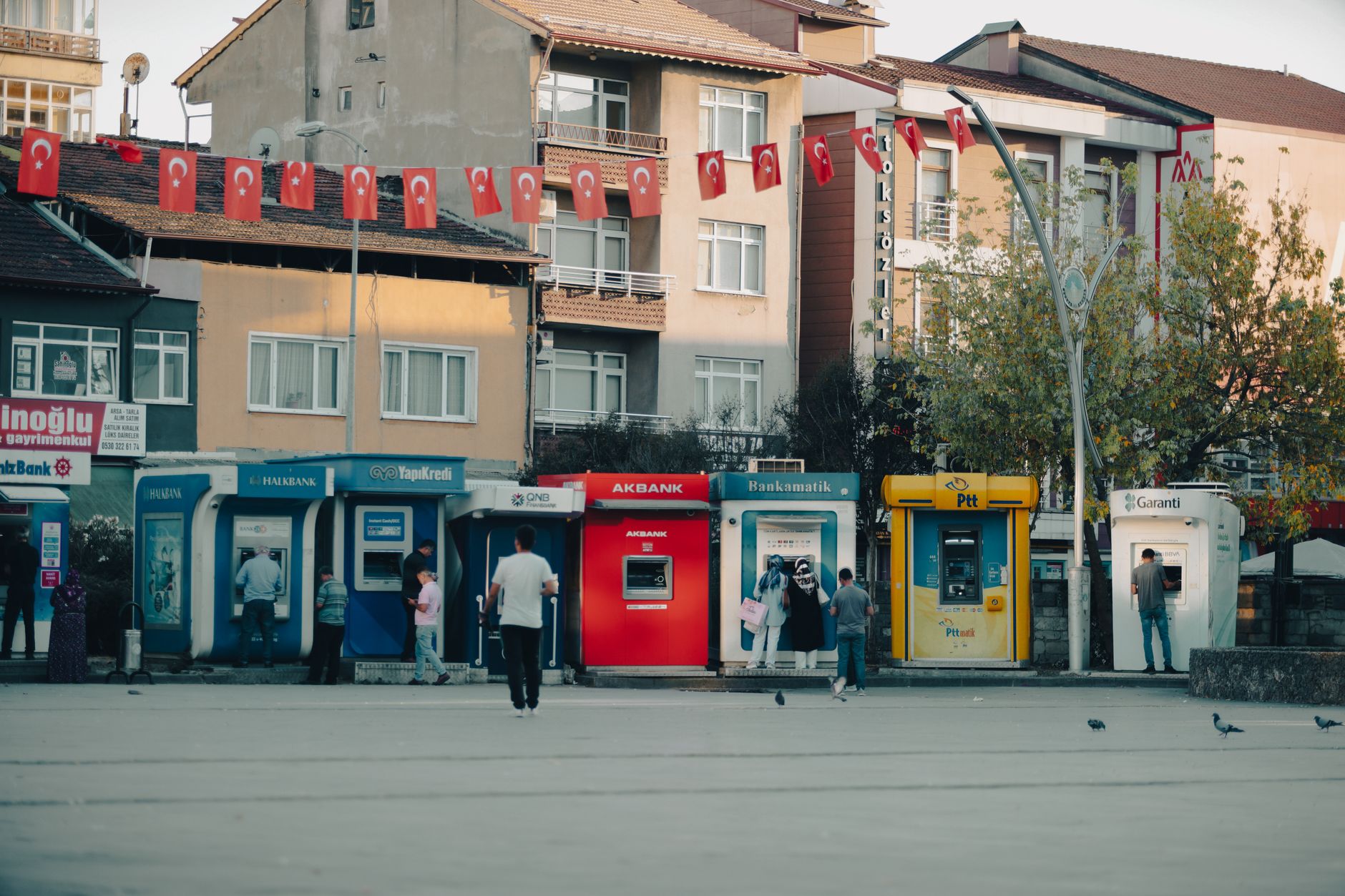 Street view in Bartın, Türkiye with ATMs, people, and waving Turkish flags.