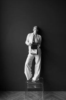 Elegant black and white portrait of a woman standing on a chair, wearing an open suit and holding a book.