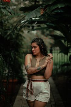 Portrait of a fashionable woman with tattoos posing under green foliage on a garden path.