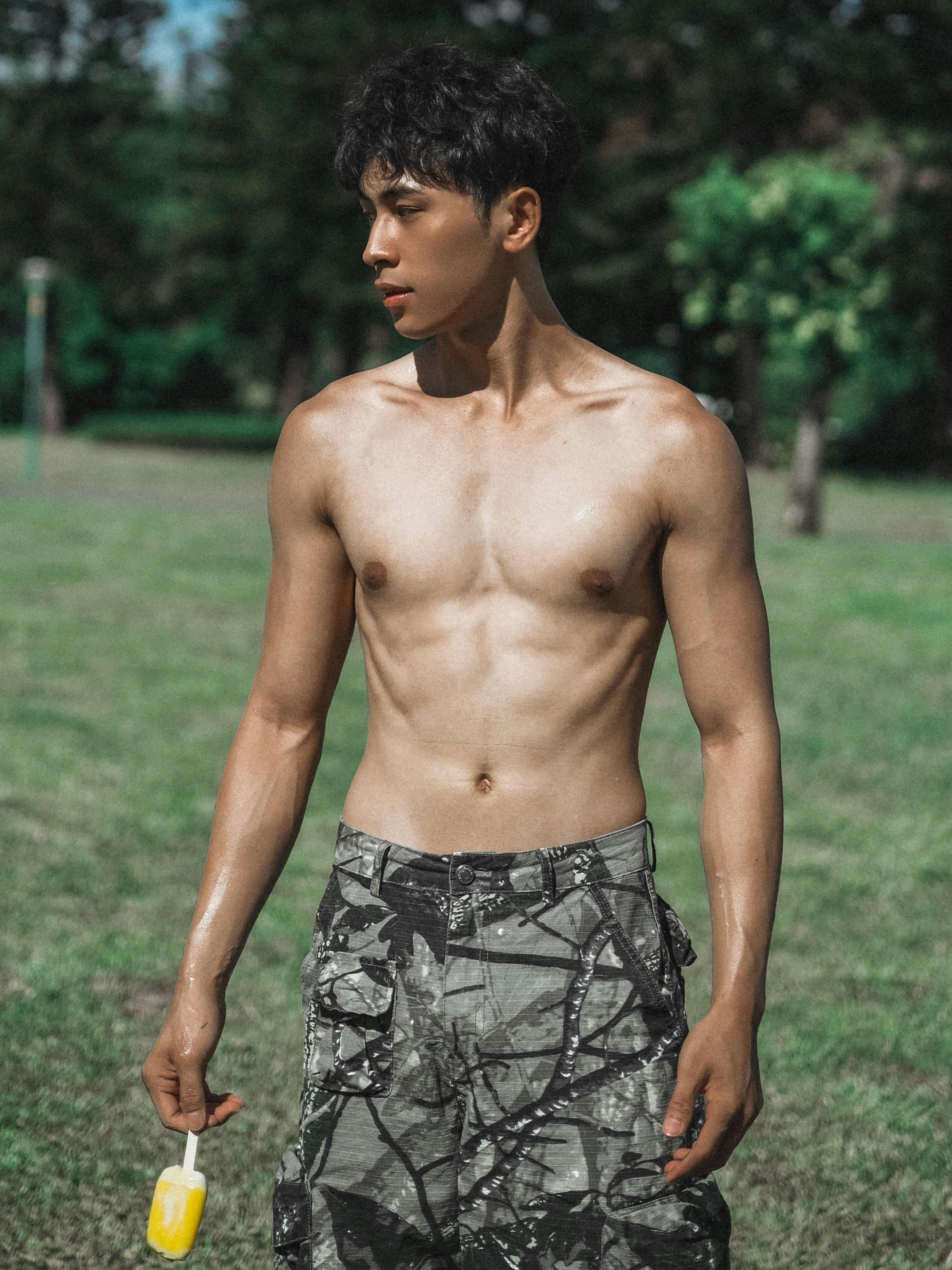 Shirtless young man holding a popsicle in a sunny park setting, epitomizing fitness and leisure.