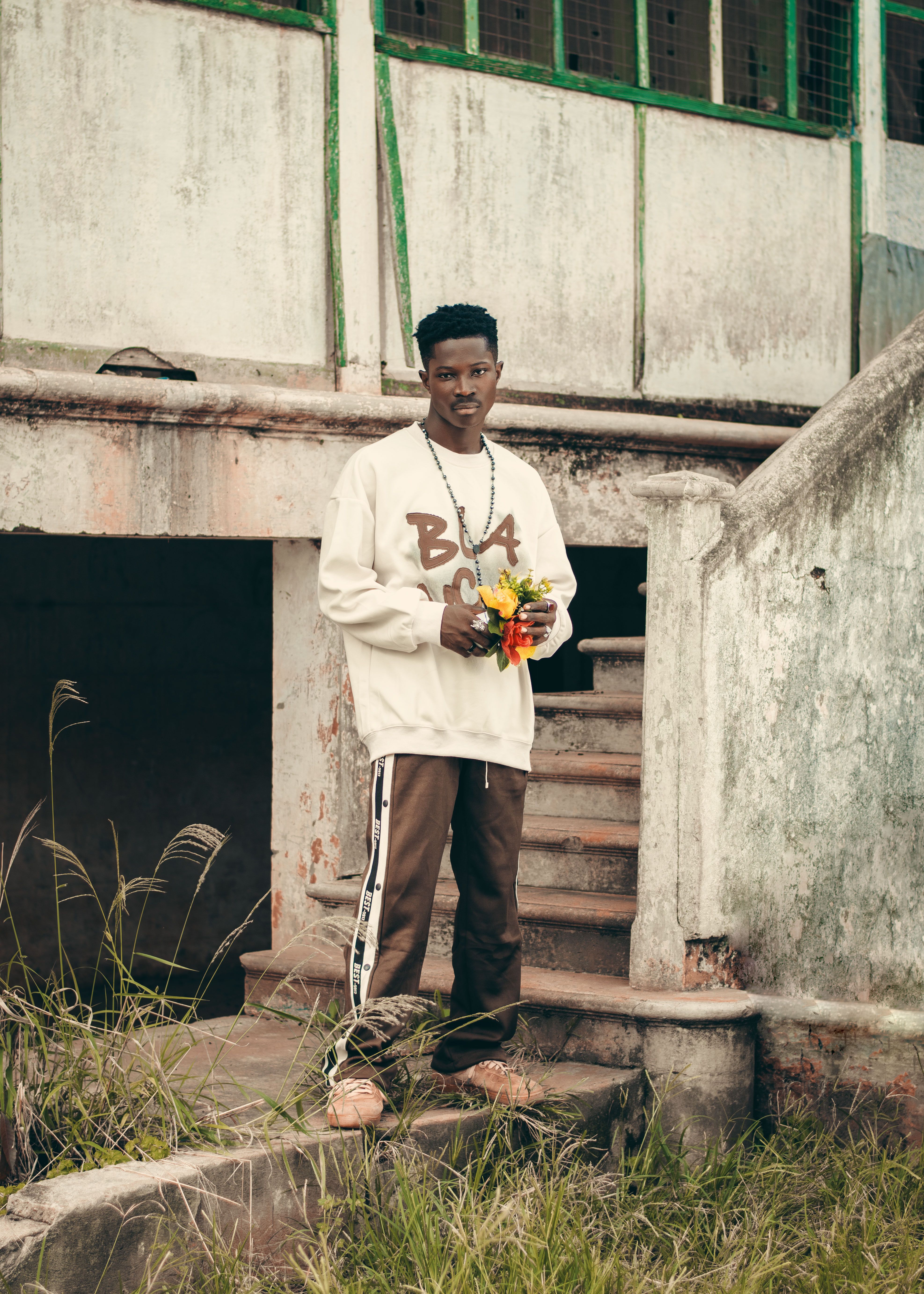 A young man stands outdoors holding flowers by an old staircase in an urban environment.