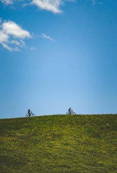 Two cyclists ride along a grassy hillside against a clear blue sky, showcasing an outdoor adventure.