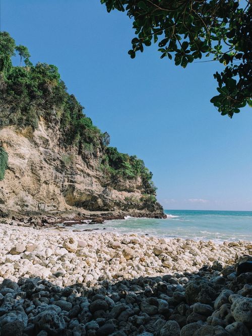 Free Relaxing beach scene with rocky shoreline, turquoise sea, and lush green cliffs under a clear blue sky. Stock Photo