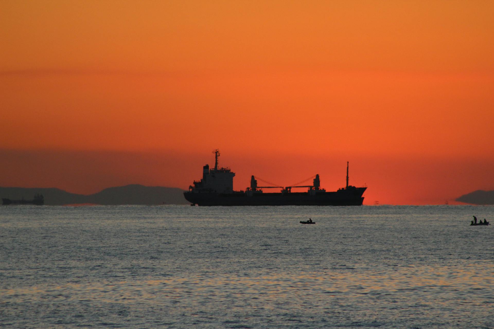 A cargo ship silhouetted against a vivid orange sunset on a calm ocean, creating a serene maritime scene.