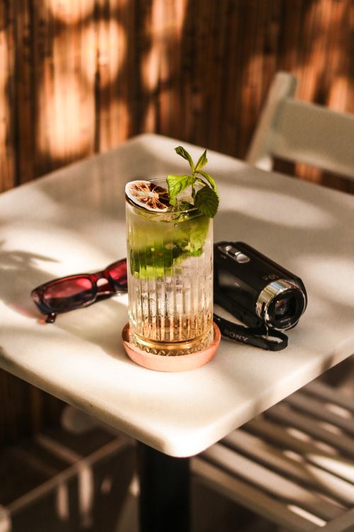 Free A chilled drink with mint and lemon on a sunny table beside a camera and sunglasses. Stock Photo