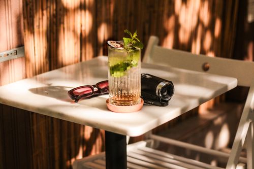 Free A mojito with mint leaves next to sunglasses and camera on a café table, warmly lit. Stock Photo
