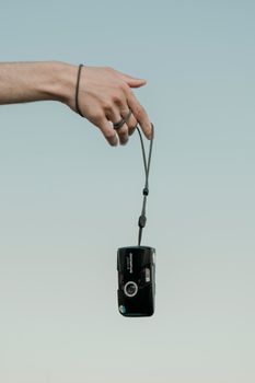 A hand holding a vintage camera with a strap, set against a clear blue sky background.